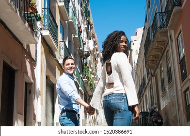 Cheerful diverse couple walking outside. Rear view of young man and woman going down old town street, holding hands and looking back. Happy tourists concept