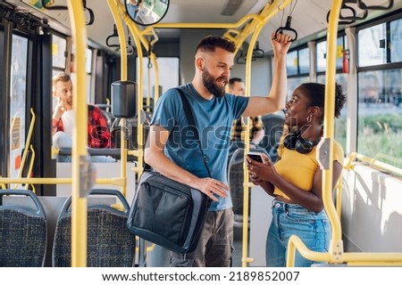 Cheerful diverse friends smiling and talking together and using a smartphone while riding a bus in the city and going to work. Standing in public transport, commuting by bus. Diverse romantic couple.
