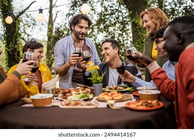 Cheerful diverse group celebrating with wine toast at alfresco dinner. Multicultural friends gathered around table with appetizers, enjoying outdoor meal in garden setting. - Powered by Shutterstock