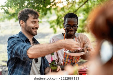 Cheerful diverse group of friends sharing food at outdoor picnic. Two men, one Caucasian and one African American, passing a plate of food, showcasing inclusivity and friendship in a natural setting. - Powered by Shutterstock