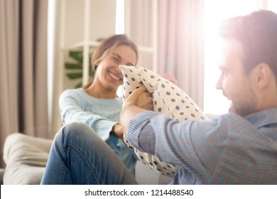 Cheerful Diverse Married Couple In Love Sitting On Sofa In Living Room At Home. Millennial Pretty Mixed Race Wife And Caucasian Husband Have Fun Playing Fighting With Pillows Laughing Enjoying Free 