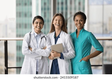 Cheerful diverse medical team of doctors standing together, looking at camera with toothy smiles, laughing. Happy young practitioner women in medic uniform coats posing for portrait - Powered by Shutterstock