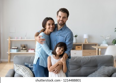 Cheerful Diverse Multi-ethnic Family Married Couple Wife Husband Little Daughter Embracing Standing Together In Living Room Smiling Looking At Camera At New Modern Home Feels Happy And Satisfied