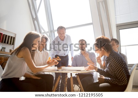 Cheerful diverse team people workers students laughing at funny joke while eating pizza together, friendly multi-ethnic colleagues group talking enjoying having fun and corporate lunch in office room