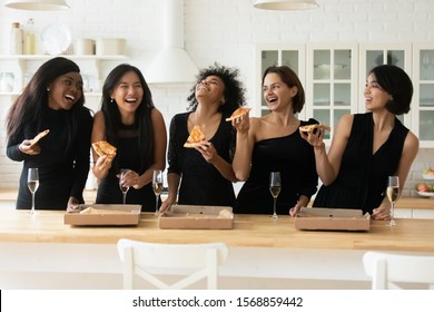Cheerful diverse young ladies laughing sharing takeaway pizza, happy funny multiethnic women wear elegant black dresses eating italian food drinking champagne having fun enjoying party in kitchen