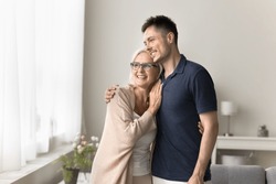 Cheerful Dreamy Adult Son And Senior Mom Standing Close At Window, Hugging With Love, Care, Looking Away, Smiling, Talking, Discussing Leisure, Family Plans, Retirement. Home Casual Portrait