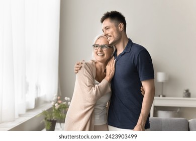 Cheerful dreamy adult son and senior mom standing close at window, hugging with love, care, looking away, smiling, talking, discussing leisure, family plans, retirement. Home casual portrait - Powered by Shutterstock