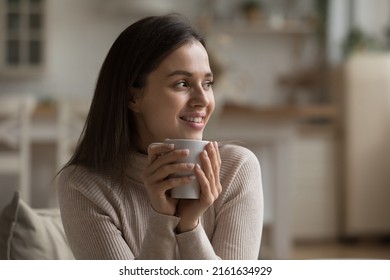 Cheerful dreamy girl holding mug of hot beverage, looking away in deep good thoughts, smiling, dreaming, enjoying aromatic herbal tea, smelling morning coffee, warming hands - Shutterstock ID 2161634929