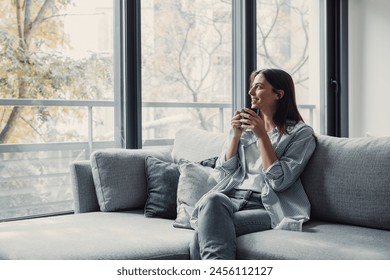 Cheerful dreamy girl holding mug of hot beverage, looking away in deep good thoughts, smiling, dreaming, enjoying aromatic herbal tea, smelling morning coffee, warming hands