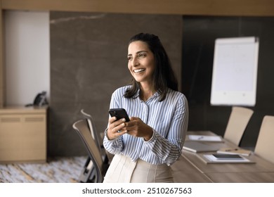 Cheerful dreamy Latin executive woman using mobile phone, digital gadget in office meeting room, holding smartphone, looking away with toothy smile, thinking on career success - Powered by Shutterstock