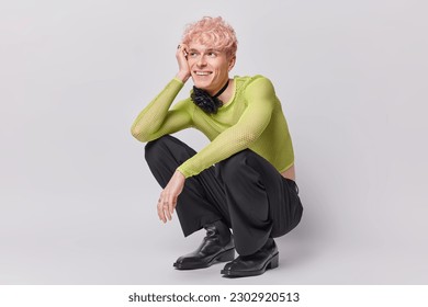 Cheerful dreamy pink haired transgender man squats indoor against white wall being in good mood recalls something pleasant in mind dressed in fashionable clothes. Non binary people and lgbt lifestyle - Powered by Shutterstock