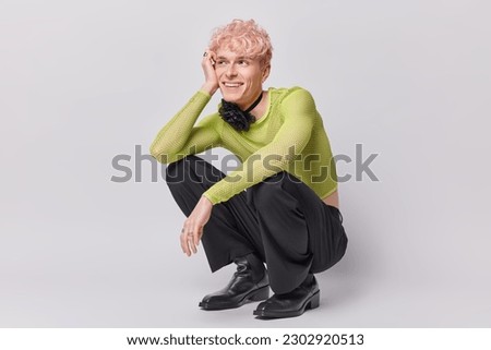 Cheerful dreamy pink haired transgender man squats indoor against white wall being in good mood recalls something pleasant in mind dressed in fashionable clothes. Non binary people and lgbt lifestyle