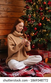 Cheerful dreamy young woman writing wish list for new year on background of Christmas tree at cozy dark living room with festive interior. Cheerful lady writing xmas letter to Santa Claus.
