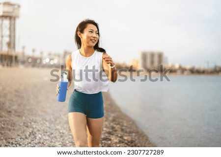 Cheerful east-Asian ethnicity young woman running on the sandy sea beach in summer sports clothing, working out