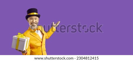 Cheerful eccentric and positive senior man man giving you gift on pastel purple background. Funny mature man in yellow stage costume and top hat holds out gift box to camera near copy space.