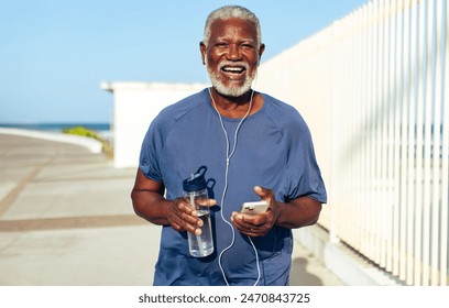 A cheerful elderly African American man listens to music on his phone while holding a water bottle during a sunny, serene walk by the sea. - Powered by Shutterstock