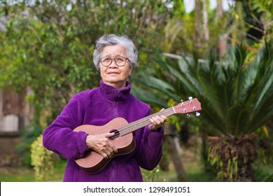 Cheerful elderly Asian woman with short gray hair wearing glasses and playing the ukulele while standing in a garden. Concept of aged people and relaxation
