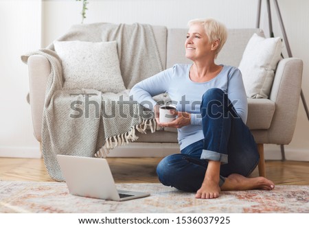 Cheerful elderly business lady sitting on floor at home, drinking coffee, using laptop, free space
