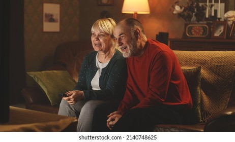 Cheerful elderly couple laughing and talking while sitting on sofa and watching funny movie on TV in weekend evening at home