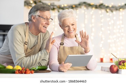 Cheerful Elderly Couple Making Video Call On Digital Tablet In Modern Kitchen Celebrating Christmas Holiday Distantly. Senior Spouses Cooking Dinner And Communicating With Remote Family Online