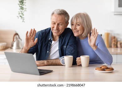 Cheerful Elderly Couple Making Video Chat With Friends While Drinking Coffee At Kitchen, Senior Man And Woman Sitting At Table, Looking At Computer Screen, Waving And Smiling, Embracing, Copy Space