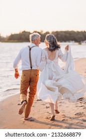 Cheerful Elderly Couple Runs Along Beach Holding Hands, Rear View.