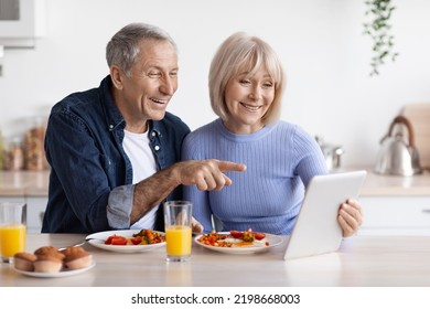 Cheerful elderly couple using digital pad while having breakfast at kitchen, elderly man and woman eating healthy food, checking photos on social media and smiling, having video conference with kids - Powered by Shutterstock