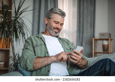 Cheerful elderly european man with beard typing on smartphone, enjoying social media chat in room interior. Social distance and new application, device for pensioner at home due covid-19 pandemic - Powered by Shutterstock