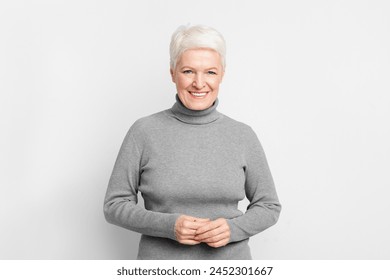 Cheerful elderly European woman with clasped hands, illustrating s3niorlife contentment on grey background - Powered by Shutterstock