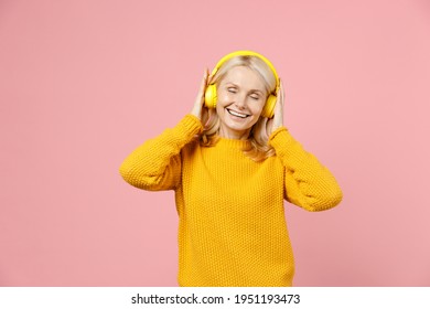 Cheerful elderly gray-haired blonde woman lady 40s 50s years old in yellow casual sweater listening music with headphones keeping eyes closed isolated on pastel pink color background studio portrait