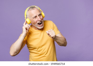 Cheerful elderly gray-haired mustache bearded man in casual yellow t-shirt posing isolated on violet wall background studio portrait. People lifestyle concept. Listen music with headphones, sing song - Powered by Shutterstock