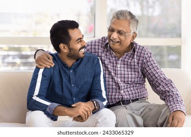 Cheerful elderly Indian granddad and happy young adult grandson talking on home couch, hugging, laughing, having fun, enjoying friendship, support, family communication - Powered by Shutterstock