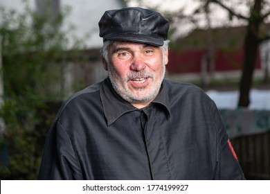 Cheerful Elderly Man With Black Old-fashioned Old Clothes And A Leather Cap Smiles And Speaks To The Camera. Looks At The Camera. Portrait On A Countryside Background. Vertical Orientation.
