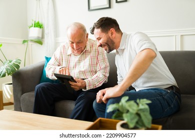 Cheerful Elderly Man Laughing While Looking At A Family Photo Frame With His Happy Son Sitting On The Sofa