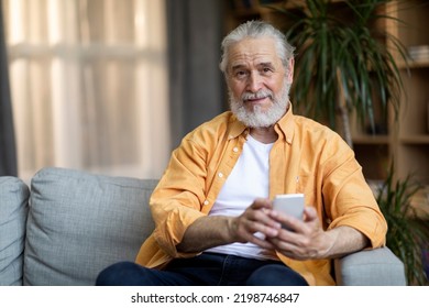 Cheerful Elderly Man Sitting On Couch At Home, Using Modern Cell Phone, Touching Gadget Screen, Smiling At Camera. Pensioner Checking Newest Entertaining Mobile App, Living Room Interior, Copy Space