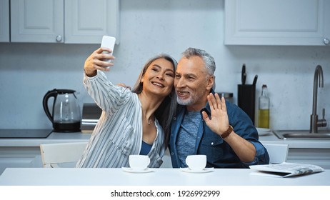 Cheerful Elderly Man Waving Hand Near Asian Wife Taking Selfie In Kitchen