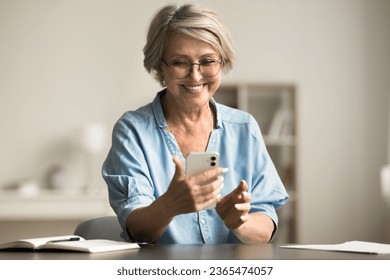 Cheerful elderly retired woman in glasses holding smartphone over home work table, smiling, laughing, using gadget for wireless online connection, reading, typing message - Powered by Shutterstock