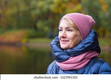 Cheerful Elderly Senior Mature Woman In Winter Hat And Scarf Smiling Walking In Golden Autumn Park With Colorful Trees. Portrait Of A Beautiful Female Pensioner Looking Into Distance. Happy Retirement