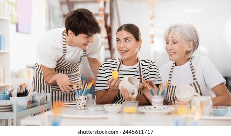 cheerful elderly woman, along with a young girl and a guy in aprons, are engaged in painting ceramic dishes at a pottery lesson. Creative hobby and the concept of communication between generations - Powered by Shutterstock