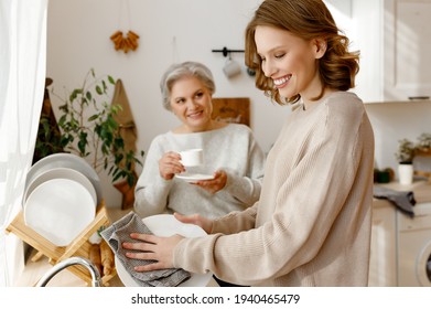 Cheerful elderly woman drinking tea and looks with a smile at her young daughter, who is washing dishes and talking   in the cozy kitchen  - Powered by Shutterstock