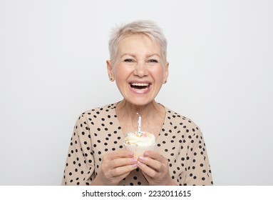 Cheerful elderly woman with short haircut holds a cupcake with a candle in her hands, celebrates a birthday and laughs, feeling happy. - Powered by Shutterstock