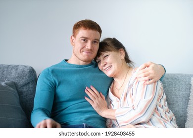 Cheerful Elderly Woman Sitting On The Sofa Next To His Adult Son. Caring Son Hugs His Elderly Mother. Relations Between Different Generations.