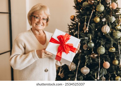 A cheerful elderly woman stands by a beautifully decorated Christmas tree, holding a wrapped gift with a red ribbon. The cozy festive atmosphere captures the spirit of holiday joy and giving. - Powered by Shutterstock