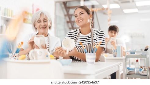 Cheerful elderly woman and young girl, participating in pottery masterclass, sitting together at table, engaging in friendly conversation while painting ceramic dishware - Powered by Shutterstock