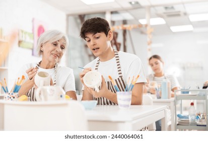 Cheerful elderly woman and young guy, participating in pottery masterclass, sitting together at table, engaging in friendly conversation while painting ceramic items.. - Powered by Shutterstock