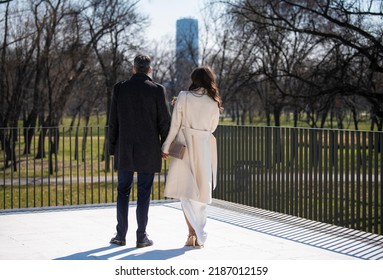 Cheerful Elegant Couple Of Bride And Groom From Behind, Standing On Terrace And Holding Hands While Looking At The Beautiful Park Scenery In Background.