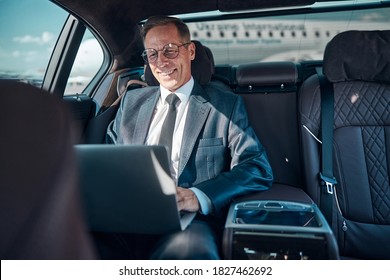 Cheerful elegant man in glasses is sitting in car with notebook while being transfered at airport - Shutterstock ID 1827462692