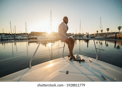 A cheerful elegant mature bald bearded black man in a summer suit consisting of striped shorts and blazer is sitting on the railings of his luxurious yacht, laughing and enjoying the dramatic sunset