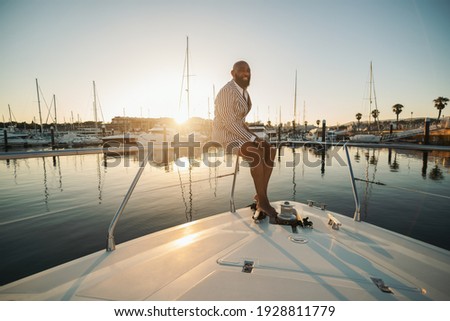 A cheerful elegant mature bald bearded black man in a summer suit consisting of striped shorts and blazer is sitting on the railings of his luxurious yacht, laughing and enjoying the dramatic sunset