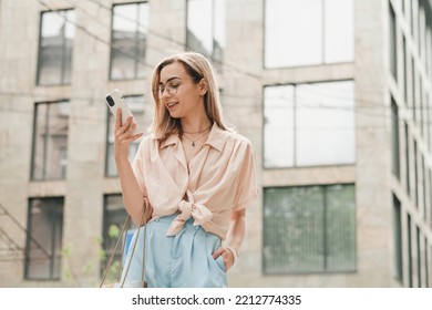 Cheerful Elegant Woman In Glasses Holding Smartphone In Hand And Smiling, Stylish Blonde Girl Outdoors Standing On The Street Looking On Cell Phone